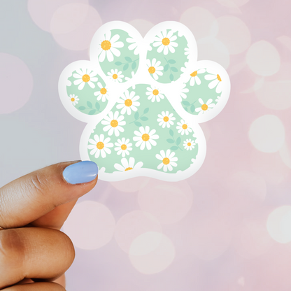 A vinyl sticker with a light green background featuring a floral pattern of daisies and a central paw print design, held by a hand against a blurred background.