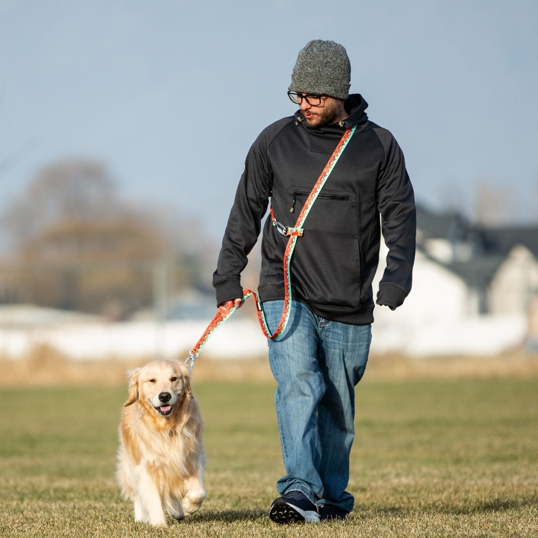 A man wearing a gray beanie, black jacket, and blue jeans walks a golden retriever on an outdoor leash in a grassy field under a clear sky.