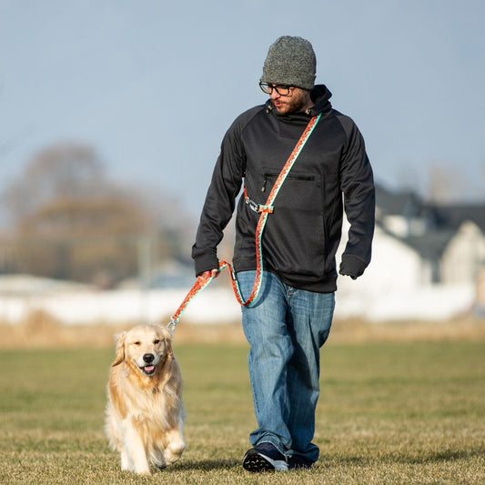 A man wearing a gray beanie, black jacket, and blue jeans walks a golden retriever on an outdoor leash in a grassy field under a clear sky.