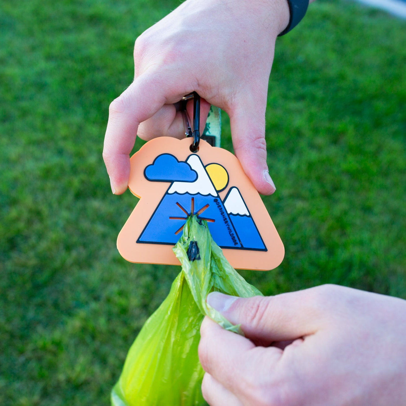 Person holding a green bag with a decorative wooden tag featuring a mountain and sun design against a grassy background.