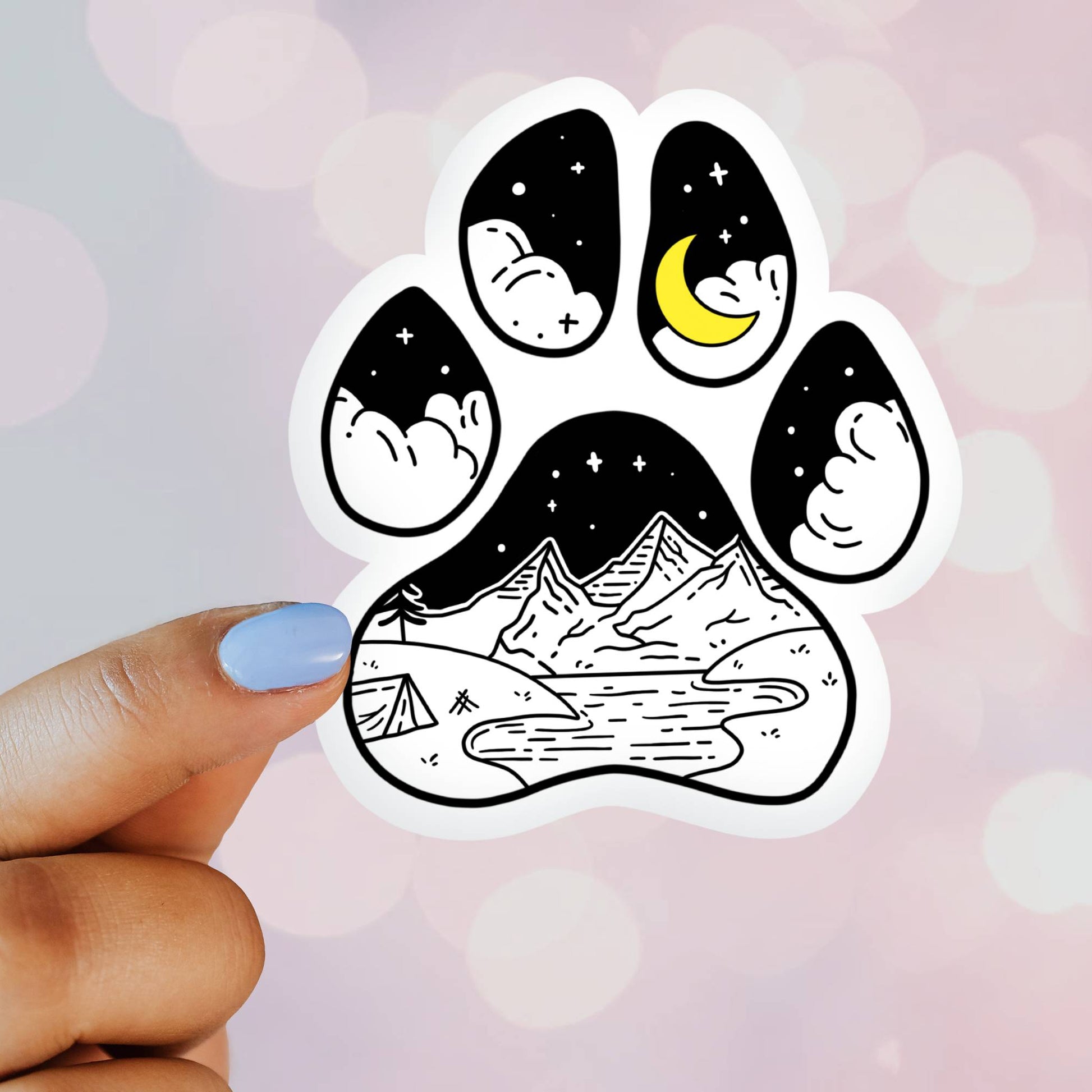 A vinyl sticker depicting a paw print with a mountain range and stars design, held between fingers against a bokeh background.